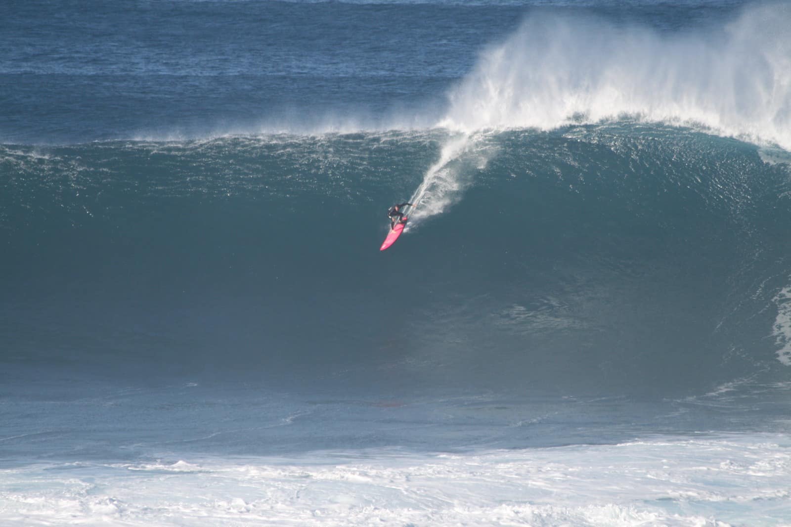 Surfista a descer uma onda grande, com espuma branca a rebentar atrás.