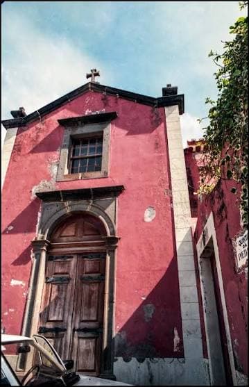Fachada rosada da capela, com portal de madeira, janela com moldura de pedra e cruz no topo.