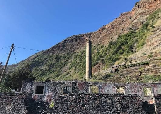 Ruínas de pedra do antigo engenho, com a chaminé alta em frente à encosta.