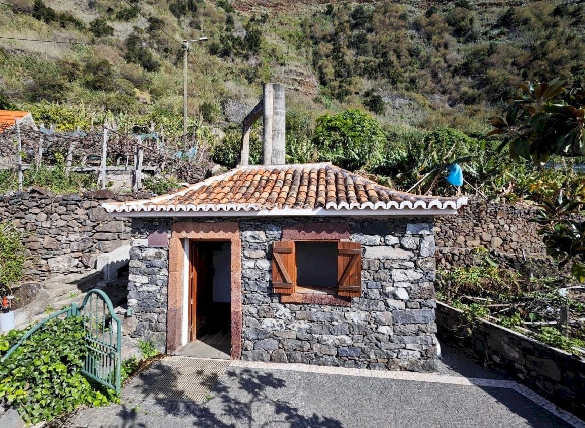 Pequena construção de pedra com telhado de telha e porta de madeira, rodeada de socalcos e bananeiras.