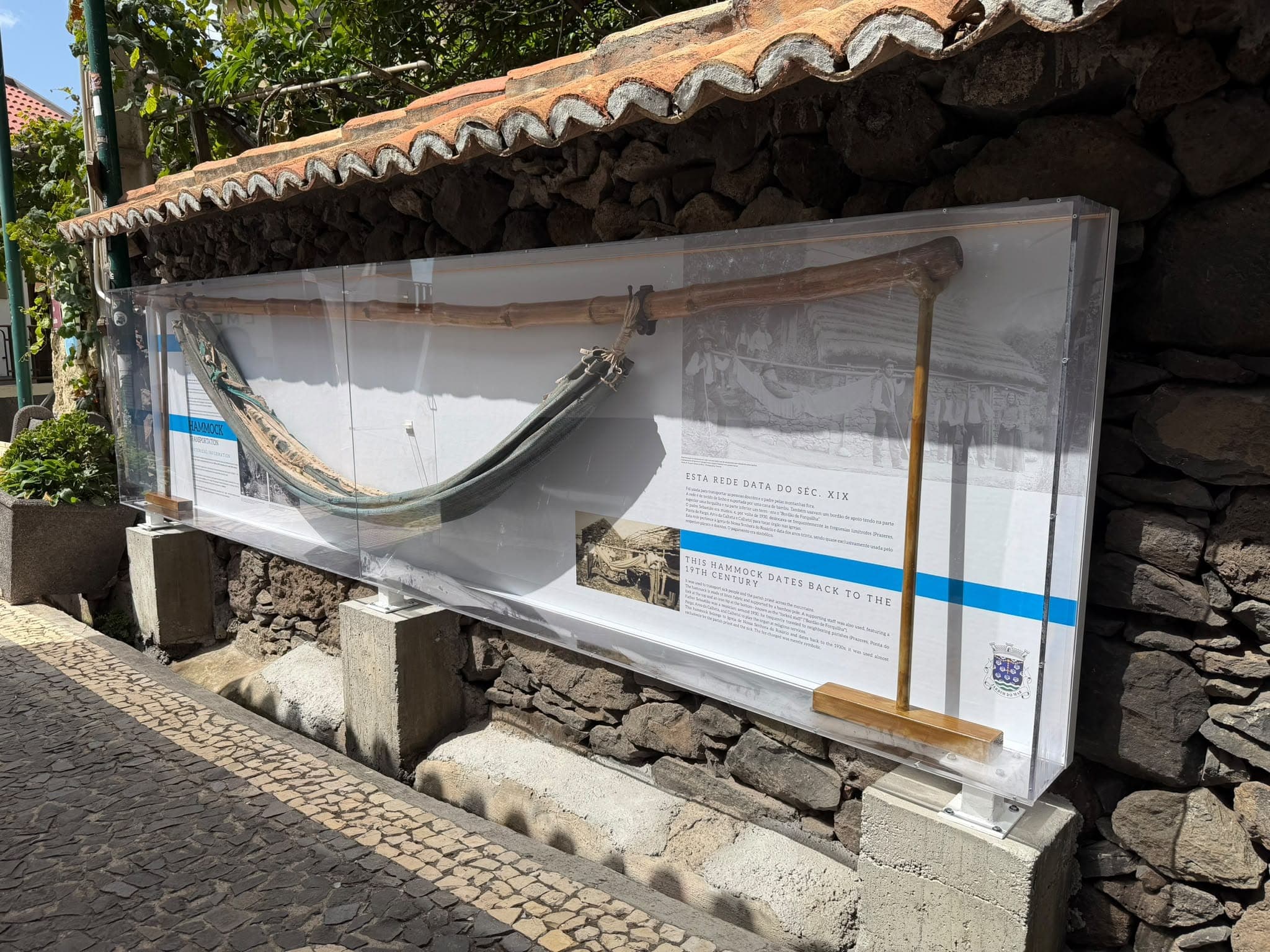 Rede de linho presa a uma cana de bambu, exposta numa vitrina junto a um muro de pedra, com painéis informativos e fotografias antigas.